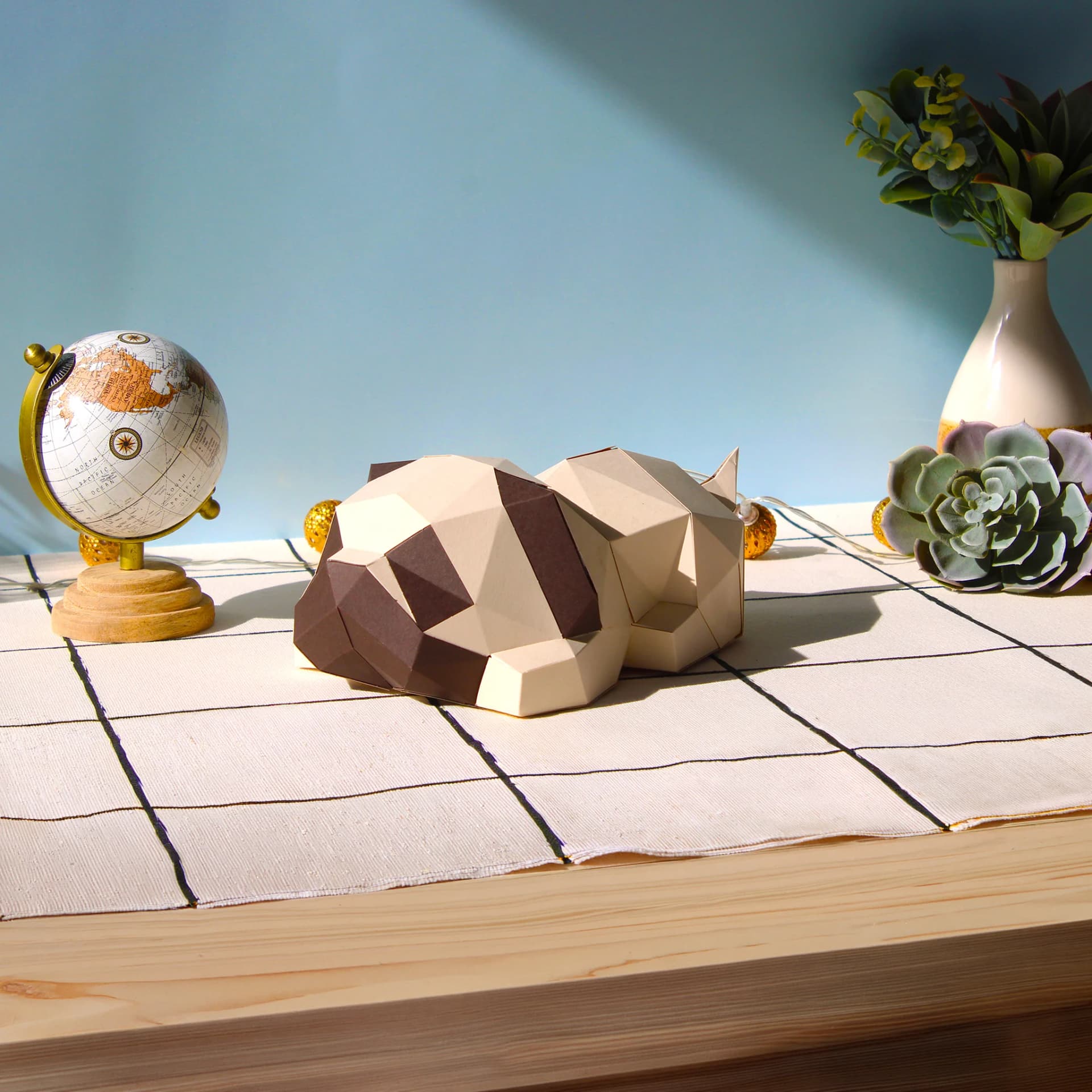 Décoration origami 3D en papier de chien carlin posé sur une commode avec une nappe a carreaux devant un mur bleu