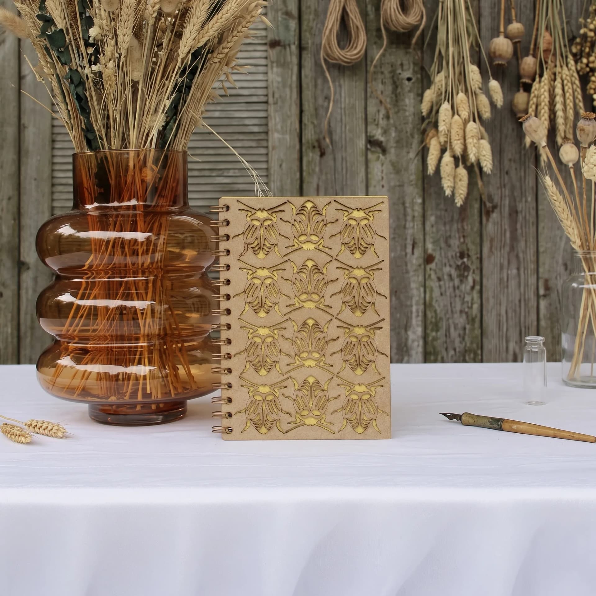 Papeterie couverture en bois de carnet insecte scarabée posé sur une table avec un vase rempli de fleurs sechés