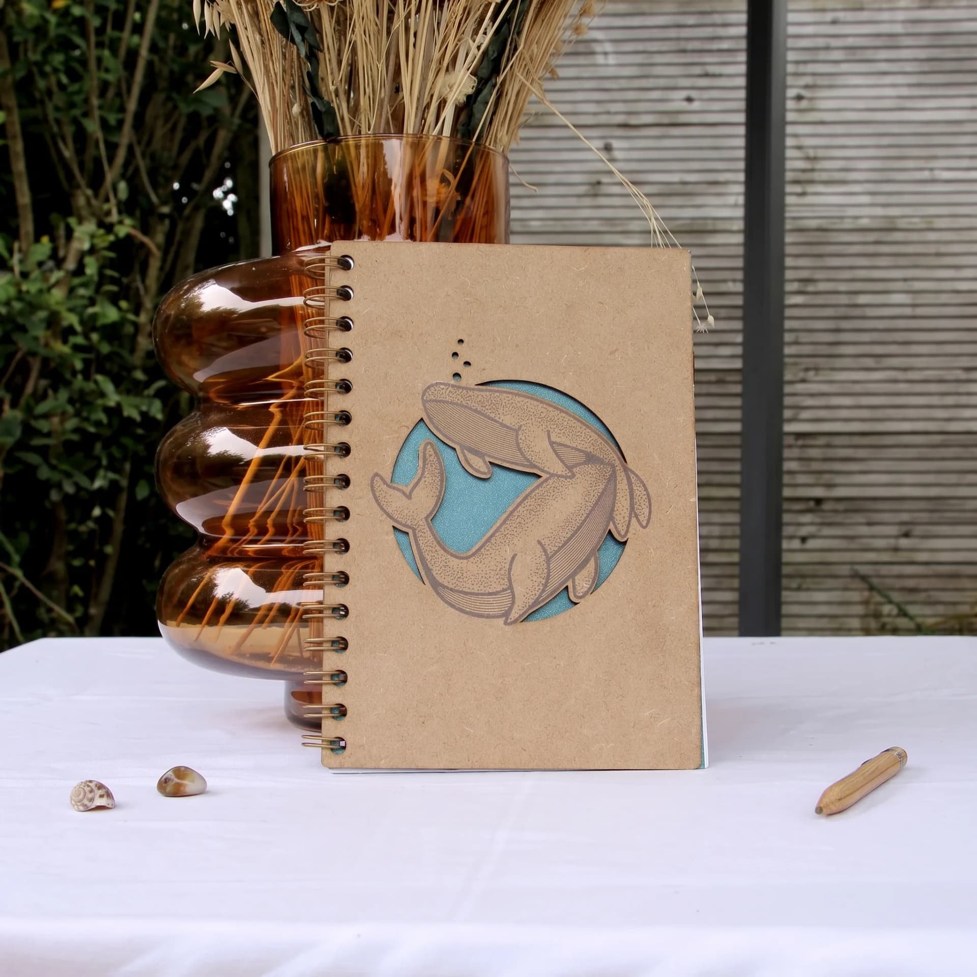 Papeterie couverture en bois de carnet marin baleine posé sur une table devant un vase rempli de fleurs sechés