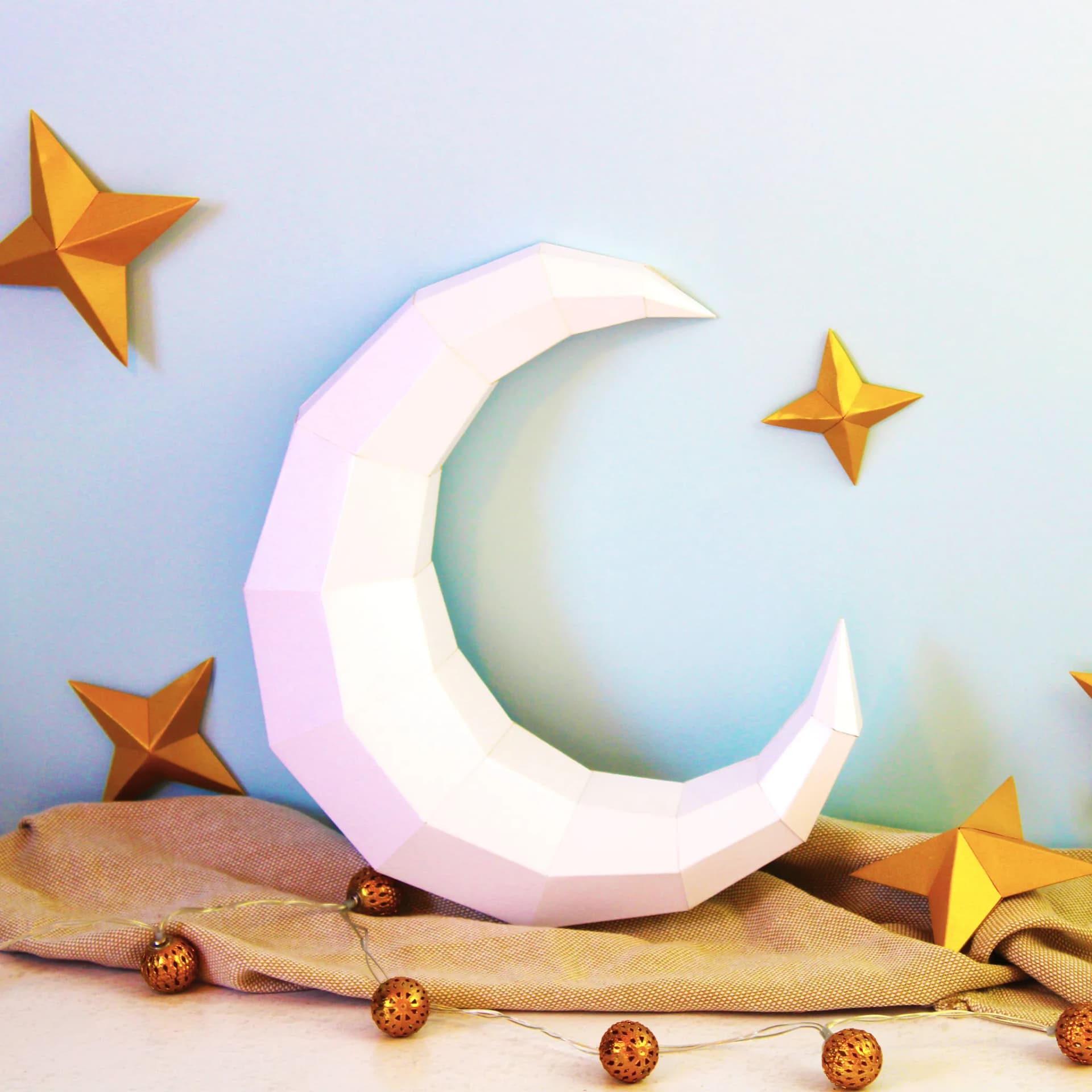 Décoration origami lune et étoiles en papier blanc et dore posées sur un tissu près d'une guirlande