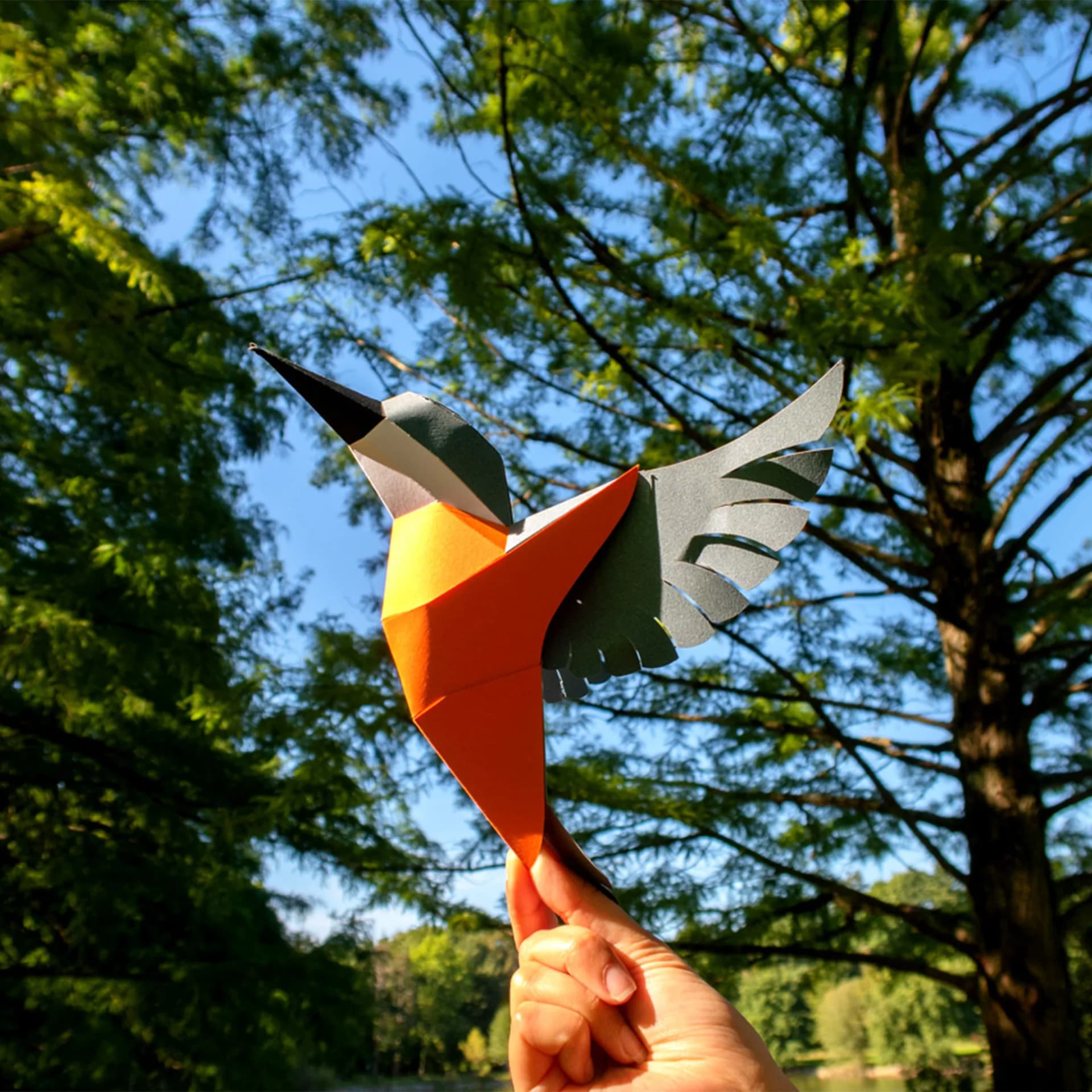 Décoration origami Martin Pêcheur en papier tenu dans une main en l'air devant des arbres